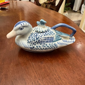 Puede incluir: Una tetera de cerámica azul y blanca con forma de pato. La tetera tiene un diseño detallado de escamas y un pequeño pato en la tapa.