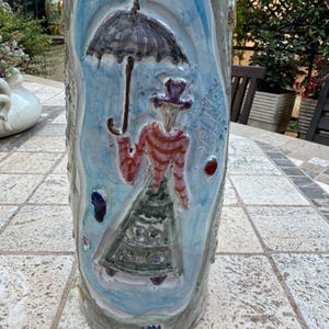 Pode incluir: Um suporte de cerâmica para guarda-chuvas com fundo azul e branco. Uma mulher com uma camisa listrada vermelha e branca e uma saia verde segura um guarda-chuva preto. O suporte é decorado com detalhes coloridos.