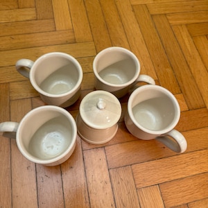 May include: Five ceramic mugs with a cream glaze and a small ceramic lidded jar. The mugs and jar are arranged on a wooden surface.
