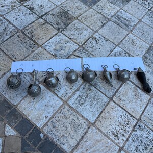 Op de afbeelding: Een verzameling van acht kleine, zilverkleurige metalen ornamenten, elk met een uniek ontwerp. De ornamenten zijn in een rij gerangschikt, met een verscheidenheid aan vormen, waaronder bollen, een vogel en een gestileerde voet. Elk heeft een kleine lus om op te hangen.