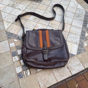May include: A brown leather crossbody bag with a striped detail on the front flap. The bag has a buckle closure and a long adjustable strap.