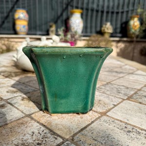 May include: A square, green ceramic planter with a glossy finish. The planter has a flared top and a slightly textured surface. Other decorative vases are visible in the background.