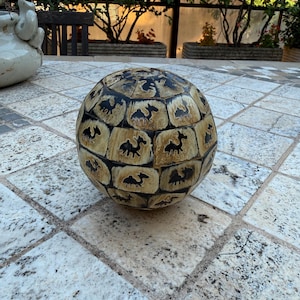 Puede incluir: Una esfera decorativa con un diseño de camello. La esfera es de color beige con detalles en marrón oscuro y un patrón repetido de siluetas de camellos. La esfera está sobre una superficie de piedra.