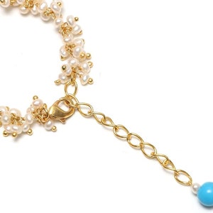 Pode incluir: Um bracelete de corrente dourada com um cacho de pérolas brancas e uma conta turquesa na ponta.