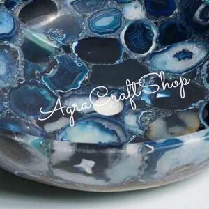 May include: A decorative, oval-shaped sink basin made of polished blue agate stone. The basin features a mosaic pattern of blue, white, and gray stone pieces. The text "Agra Craft Shop" is written across the front of the basin.