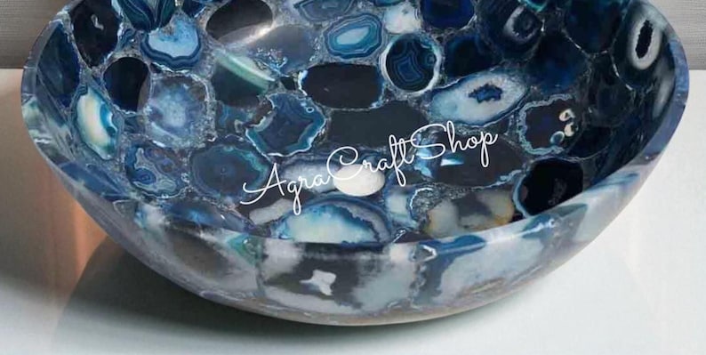 May include: A decorative, oval-shaped sink basin made of polished blue agate stone. The basin features a mosaic pattern of blue, white, and gray stone pieces. The text "Agra Craft Shop" is written across the front of the basin.