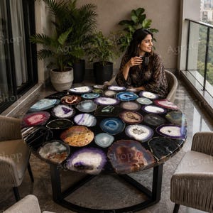 May include: A circular table featuring a black surface adorned with numerous agate slices in shades of purple, brown, and blue. The table is supported by a black metal base and is surrounded by two neutral-toned chairs. Plants and a woman are in the background.
