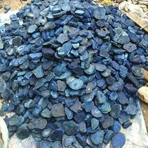 May include: A large pile of raw, uncut blue agate stones. The stones vary in size and shape, with a mix of dark and light blue hues. Some stones have visible banding and natural patterns. The pile is resting on a light-colored surface.