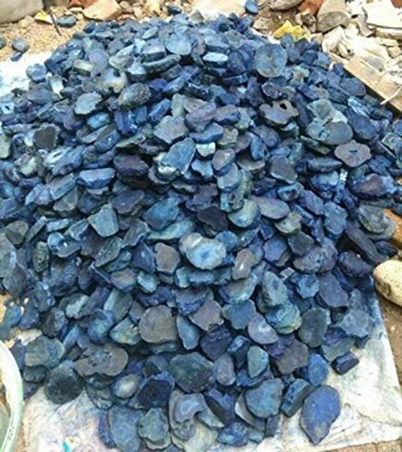May include: A large pile of raw, uncut blue agate stones. The stones vary in size and shape, with a mix of dark and light blue hues. Some stones have visible banding and natural patterns. The pile is resting on a light-colored surface.