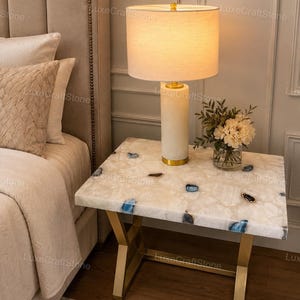 May include: A rectangular side table with a white stone top featuring embedded blue and brown agate accents, supported by a gold-toned metal frame. A cylindrical lamp with a white fabric shade and a gold base sits on the table next to a glass vase.