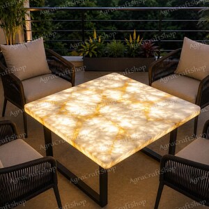 Peut inclure: Une table carrée en pierre d'onyx rétroéclairée avec un motif marbré blanc et ambre lumineux, soutenue par un cadre en métal noir, entourée de quatre chaises tressées marron foncé avec des coussins beige clair sur un balcon.