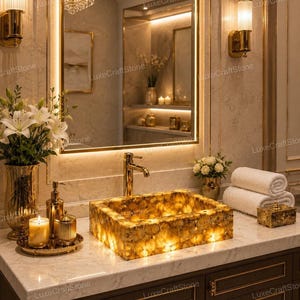 May include: A rectangular, backlit amber-toned stone sink sits on a white marble countertop. The sink features a warm, glowing internal light. A gold faucet, a gold soap dispenser, a candle, and white towels are arranged on the vanity surface.