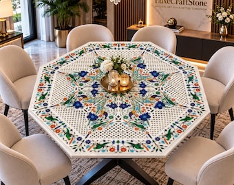 May include: An octagonal white stone dining table featuring an intricate inlaid floral pattern in blue, green, and orange. The table has a black geometric base and is surrounded by six beige upholstered chairs with dark wooden legs.