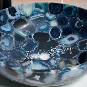 May include: A handcrafted, round bowl with a mosaic design. The bowl is made of polished, blue-toned agate stones. The text "Agra Craft Shop" is written in cursive across the bowl's surface. The bowl's interior is a deep blue.
