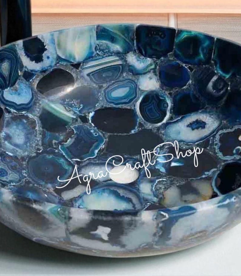 May include: A handcrafted, round bowl with a mosaic design. The bowl is made of polished, blue-toned agate stones. The text "Agra Craft Shop" is written in cursive across the bowl's surface. The bowl's interior is a deep blue.