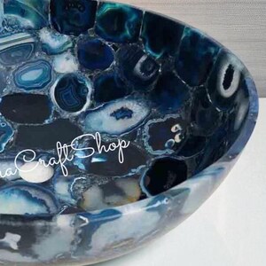 May include: A handcrafted, oval-shaped sink basin made of polished blue agate stone. The basin features a mosaic pattern of various shades of blue, white, and gray, with the text "Craft Shop" in cursive script.