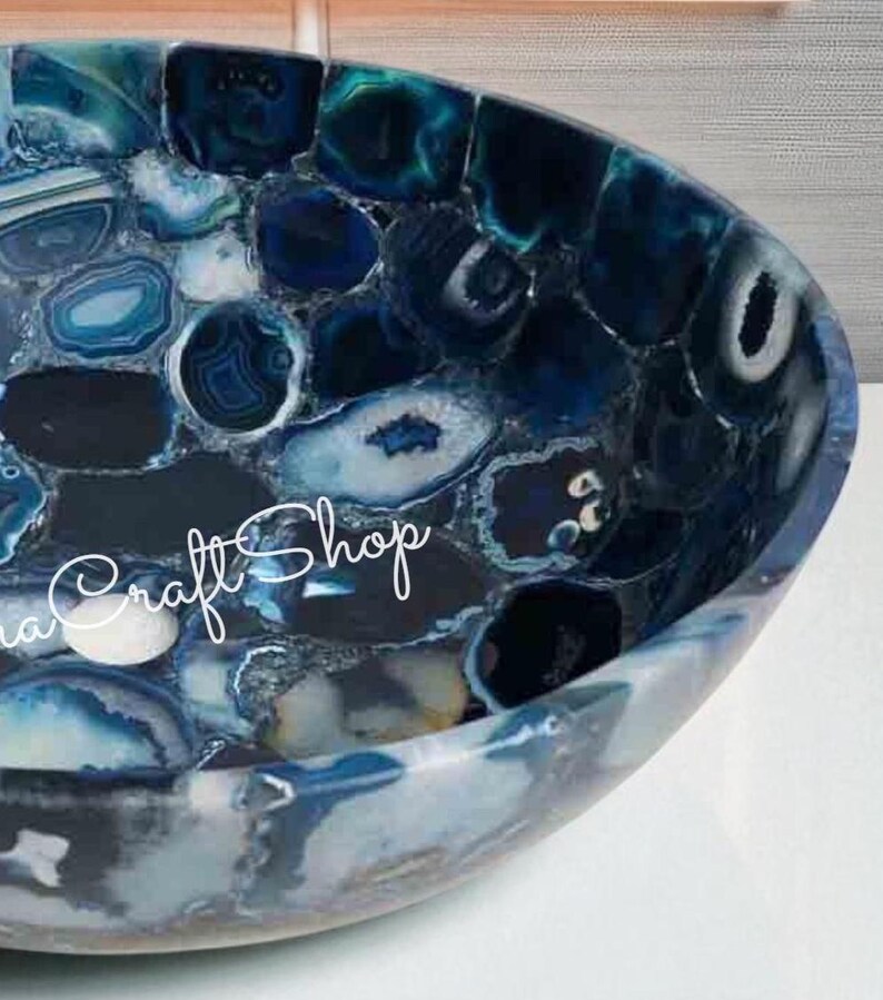 May include: A handcrafted, oval-shaped sink basin made of polished blue agate stone. The basin features a mosaic pattern of various shades of blue, white, and gray, with the text "Craft Shop" in cursive script.