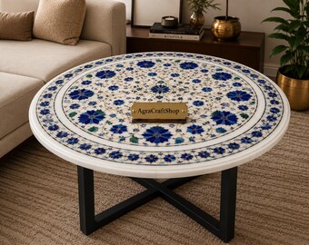 May include: A round white marble coffee table featuring an intricate inlay of blue and green floral patterns. The tabletop is supported by a black metal X-shaped base. A small rectangular brass plaque with the text AgraCraftShop is centred.