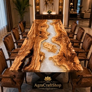 Op de afbeelding: Een rechthoekige eettafel met een olijfhouten blad met natuurlijke randen en een centrale inleg van doorschijnend wit hars, rustend op een zwart metalen onderstel. De tafel is omringd door acht bruine gestoffeerde stoelen.
