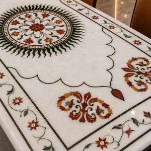 May include: A rectangular white marble tabletop featuring intricate floral inlay work. The design includes a central sunburst motif with red and orange flowers, surrounded by scrolling vine borders and symmetrical leaf patterns in red, orange, and green.