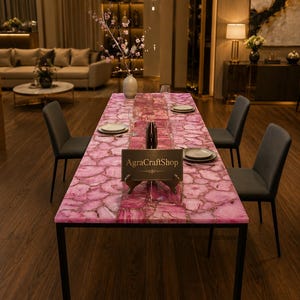 May include: A rectangular dining table featuring a pink agate stone tabletop with a natural geode pattern and gold-toned veining, supported by a black metal frame. A small black sign with the gold text AgraCraftShop sits on the table surface.