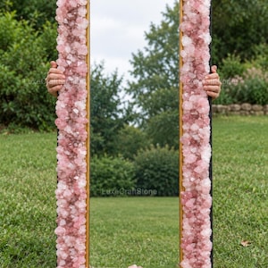 May include: A rectangular mirror featuring a thick frame covered in numerous raw, light pink rose quartz crystals. The mirror has a thin gold-colored inner border and a black outer edge, reflecting a green outdoor landscape with trees and grass.