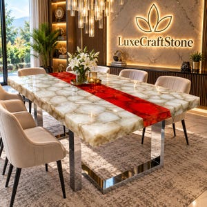 Puede incluir: Una mesa de comedor rectangular con una superficie de piedra blanca translúcida y una incrustación central de piedra roja. La mesa está sostenida por un marco de metal cromado pulido y rodeada por seis sillas tapizadas en color beige.