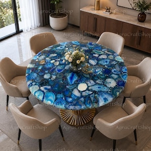 May include: A round dining table featuring a tabletop composed of polished blue agate slices in varying shades of azure, cobalt, and white, supported by a fluted gold-toned metal pedestal base, surrounded by six beige upholstered chairs.