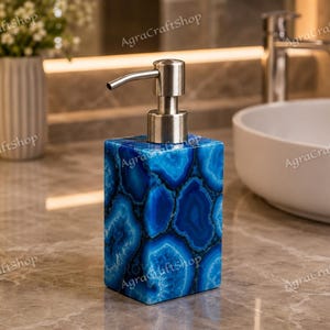 Puede incluir: Un dispensador de jabón rectangular con un patrón de piedra de ágata azul vibrante y acabado pulido. El dispensador está rematado con un mecanismo de bomba de metal plateado cepillado. El texto AgraCraftShop es visible en la imagen.