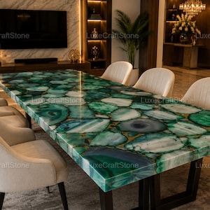Può includere: Un tavolo da pranzo rettangolare con una superficie lucida composta da fette di pietra agata verde intarsiate con varie tonalità smeraldo e verde acqua, sostenuto da una struttura in metallo nero e circondato da sei sedie imbottite color crema.