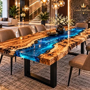 Puede incluir: Una mesa de comedor rectangular con un tablero de madera de borde natural y un río central de resina epoxi azul translúcida. La mesa se apoya sobre patas de metal negro en forma de U y está rodeada por seis sillas tapizadas en color topo.