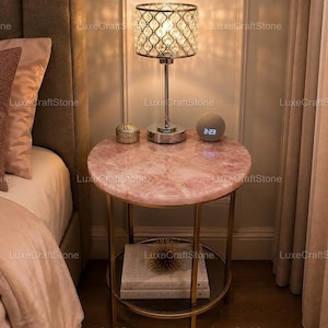 Puede incluir: Una mesa auxiliar redonda con una superficie de cuarzo rosa pulido y una estructura de metal dorado. La mesa cuenta con un estante inferior de cristal que sostiene dos libros apilados y una esfera decorativa dorada. Una lámpara de cristal y un reloj.