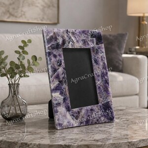 May include: A rectangular picture frame crafted from polished purple amethyst stone pieces. The frame features a natural crystalline texture with varying shades of deep violet and white veining, surrounding a solid black rectangular center insert.