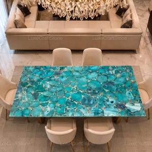 Puede incluir: Una mesa de comedor rectangular con una superficie pulida hecha de numerosas rodajas de piedra ágata azul turquesa interconectadas, apoyada sobre una base y rodeada por seis sillas tapizadas en color crema con patas de metal dorado.