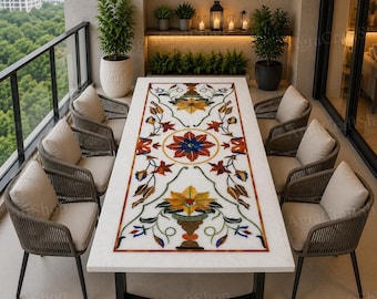 May include: A rectangular white marble table with a floral inlay design in red, blue, yellow, and green. Six woven chairs with beige cushions surround the table. The setting is on a balcony with a view of trees and plants.
