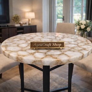 May include: A round table featuring a white and tan translucent stone top with a cracked, mosaic-like pattern, supported by a black metal base. A rectangular dark brown sign with gold trim and the text AgraCraft Shop sits on the table surface.