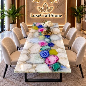 May include: A rectangular dining table featuring a polished surface made of multicolored agate slices in shades of pink, blue, green, and yellow, set against white stone. The table has a black metal base and is surrounded by eight beige chairs.
