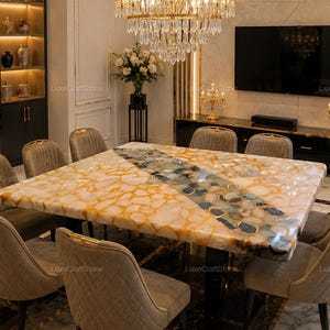 May include: A square dining table featuring a polished surface made of amber-toned stone fragments with a central diagonal inlay of blue and grey agate slices, supported by a black base and surrounded by eight beige upholstered dining chairs.