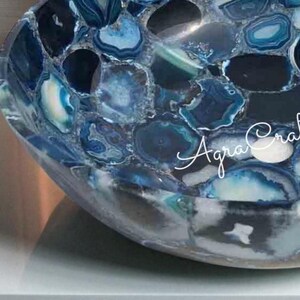 May include: A handcrafted, blue agate stone bowl with a mosaic design. The bowl features a variety of blue shades, from deep navy to light turquoise, creating a unique pattern. The text "Agra Craft" is visible on the side of the bowl.