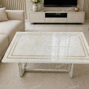May include: Rectangular white coffee table with a gold trim and silver legs. The table has a glossy finish and is placed on a neutral-colored rug. The table is a modern piece of furniture.