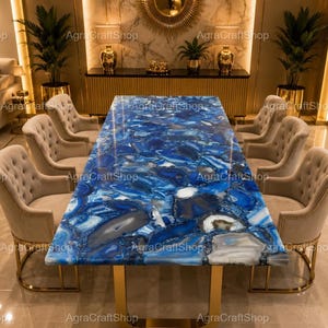 May include: A rectangular dining table featuring a polished blue agate gemstone surface with varied shades of blue, white, and grey, supported by a gold metal frame, surrounded by six beige tufted upholstered chairs with gold metal bases.