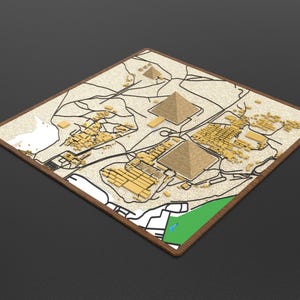 May include: A square, flat map display featuring a textured, sandy-colored surface with raised, three-dimensional golden pyramid structures and architectural blocks. The map includes black line work depicting paths, white and green zones, and a dark brown border.
