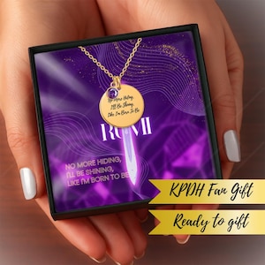 Op de afbeelding: Een goudkleurige hangerketting met een ronde bedel en een kleine paarse edelsteen. De bedel heeft de tekst "No more Hiding, I'll Be Shining, Like I'm Born To Be" en de naam "RUMI". De ketting zit in een zwarte geschenkdoos met paarse accenten.