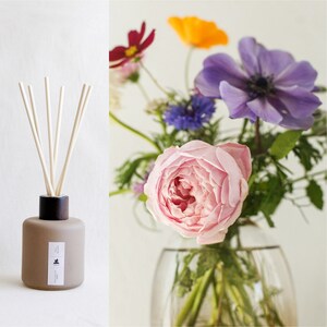 May include: A brown glass diffuser bottle with wooden sticks and a label that says "H&K". The bottle is sitting on a white surface. The background is a bouquet of flowers in a clear glass vase. The flowers include pink roses, purple anemones, orange and yellow flowers, and green foliage.