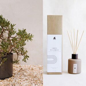 May include: A brown cardboard box with the text "GREEN TEA GROVE" and a small plant illustration. The box is next to a brown glass diffuser bottle with wooden sticks.