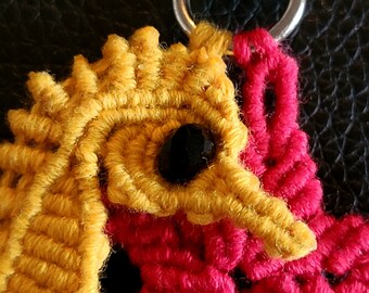 May include: A close-up of a yellow and red crocheted keychain with a black bead for an eye. The keychain is attached to a silver metal clasp.