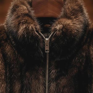 May include: A close-up shot of a brown fur coat featuring a high collar and a silver zipper. The fur has a soft, luxurious appearance with different shades of brown. The central zipper is a key design element.