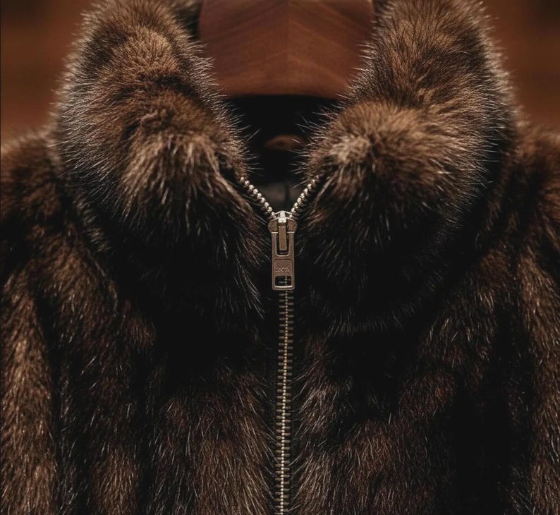 May include: A close-up shot of a brown fur coat featuring a high collar and a silver zipper. The fur has a soft, luxurious appearance with different shades of brown. The central zipper is a key design element.