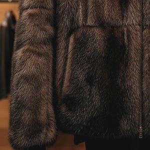 May include: A detailed view of a brown fur jacket featuring a zip fastening. The jacket's texture is dense and luxurious, with individual fur fibres clearly visible. The sleeves and front are in focus, highlighting the garment's rich colour and detail.