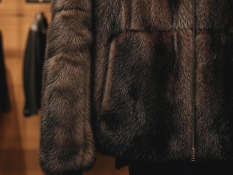 May include: A detailed view of a brown fur jacket featuring a zip fastening. The jacket's texture is dense and luxurious, with individual fur fibres clearly visible. The sleeves and front are in focus, highlighting the garment's rich colour and detail.